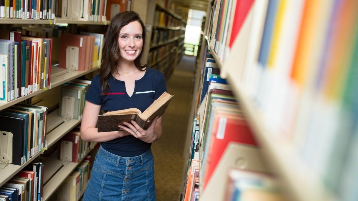 Young woman holds a book while standing among the stacks of the Coates Library